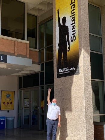 A man stands outside holding his hand in a pitchfork gesture, mimicking an ASU marketing sign above him.